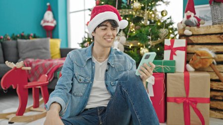 Foto de Joven hombre hispano usando smartphone celebrando la Navidad en casa - Imagen libre de derechos