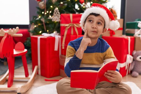 Photo for Adorable hispanic boy reading book sitting on floor by christmas tree at home - Royalty Free Image