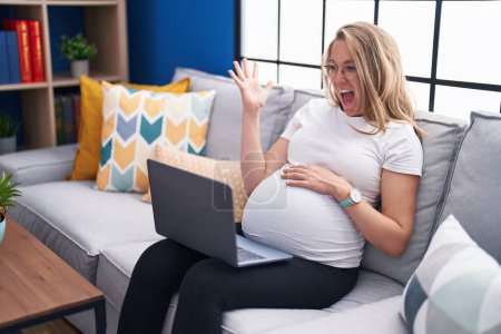 Photo for Young pregnant woman sitting on the sofa at home using laptop celebrating victory with happy smile and winner expression with raised hands - Royalty Free Image