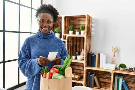 Téléchargez les photos : Femme afro-américaine souriante confiante en lisant les reçus alimentaires à la maison - en image libre de droit