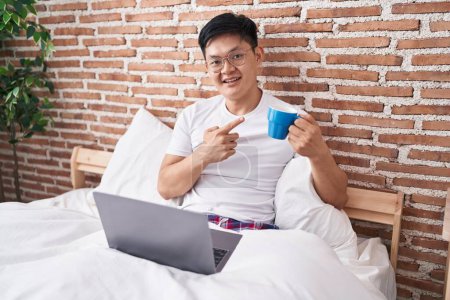Téléchargez les photos : Jeune homme asiatique boire du café assis sur le lit souriant heureux pointant avec la main et le doigt - en image libre de droit