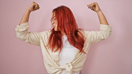 Photo for Joyful young redhead sportswoman confidently flexing arms, flaunting power & happiness over isolated pink background, a portrait of cheerful, sporty confidence. - Royalty Free Image