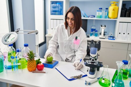 Téléchargez les photos : Jeune belle hispanique femme scientifique pesant pomme écriture sur document au laboratoire - en image libre de droit