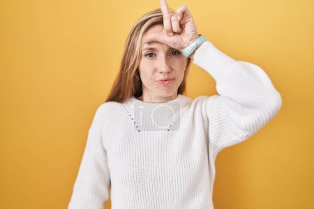 Photo for Young caucasian woman wearing white sweater over yellow background making fun of people with fingers on forehead doing loser gesture mocking and insulting. - Royalty Free Image