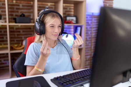 Téléchargez les photos : Jeune femme caucasienne jouant à des jeux vidéo portant des écouteurs faisant geste d'argent avec les mains, demandant le paiement du salaire, entreprise millionnaire - en image libre de droit