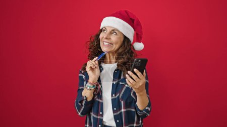 Téléchargez les photos : Moyen Age hispanique femme shopping avec smartphone et carte de crédit portant chapeau de Noël sur fond rouge isolé - en image libre de droit