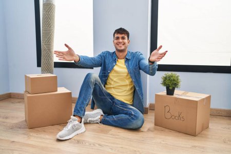 Photo for Young hispanic man sitting on the floor at new home looking at the camera smiling with open arms for hug. cheerful expression embracing happiness. - Royalty Free Image