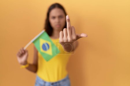 Foto de Young hispanic woman holding brazil flag showing middle finger, impolite and rude fuck off expression - Imagen libre de derechos