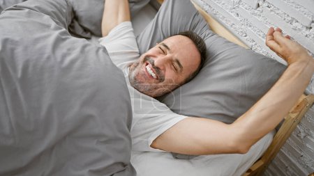 Photo for Beaming middle-aged man with grey hair joyfully stretching arms waking up in his cozy bedroom bed, relishing the comfort of a relaxing morning indoors - Royalty Free Image