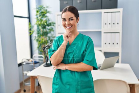 Foto de Joven hermosa enfermera hispana sonriendo confiada de pie con los brazos cruzados gesto en la clínica - Imagen libre de derechos