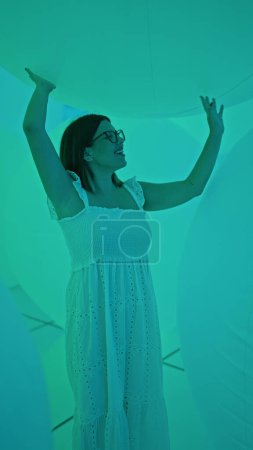 Foto de Hermosa mujer hispana con gafas disfruta de la exhibición de globos iluminados por neón en un museo futurista y moderno - Imagen libre de derechos