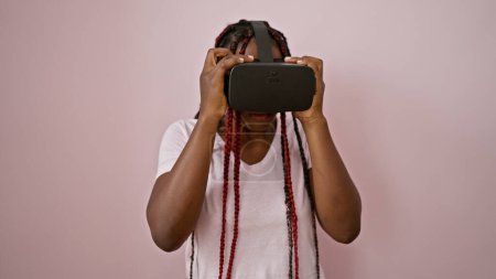 Photo for Beautiful african american woman, a serious gamer, immersed in an online game using vr glasses, standing against an isolated pink background in a relaxed, concentrated stance - Royalty Free Image