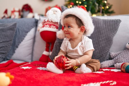 Foto de Adorable bebé hispano sosteniendo la bola de decoración de Navidad sentado en el sofá en casa - Imagen libre de derechos