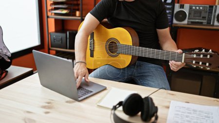 Foto de Joven músico hispano tocando guitarra clásica usando laptop en estudio de música - Imagen libre de derechos