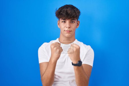 Téléchargez les photos : Adolescent hispanique debout sur fond bleu prêt à se battre avec geste de défense poing, visage en colère et bouleversé, peur du problème - en image libre de droit