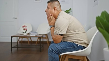 Photo for Stressed young latin man alone and frustrated, sitting in a chair, tormented by his problems, exudes an aura of sadness in the sterile indoor waiting room - Royalty Free Image