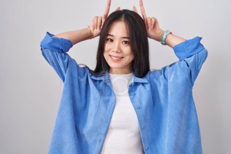 Téléchargez les photos : Jeune femme chinoise debout sur fond blanc posant drôle et fou avec les doigts sur la tête comme des oreilles de lapin, souriant gai - en image libre de droit
