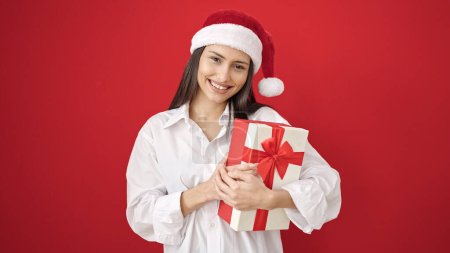 Foto de Joven hermosa mujer hispana sonriendo confiada sosteniendo regalo de Navidad sobre fondo rojo aislado - Imagen libre de derechos