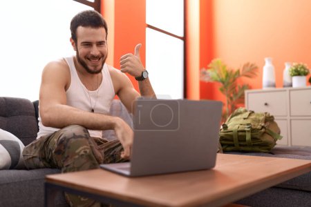 Téléchargez les photos : Jeune homme hispanique portant un uniforme de camouflage de l'armée faisant appel vidéo à la maison souriant heureux et positif, pouce levé faire excellent et signe d'approbation - en image libre de droit
