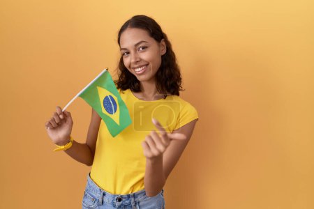 Photo for Young hispanic woman holding brazil flag beckoning come here gesture with hand inviting welcoming happy and smiling - Royalty Free Image