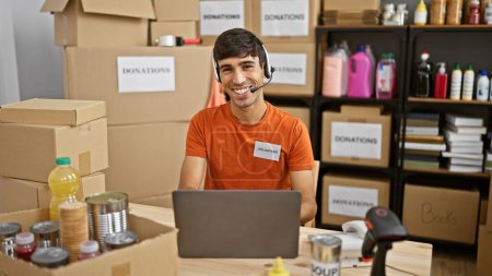 Téléchargez les photos : Beau jeune homme hispanique bénévolement, travaillant sur un ordinateur portable avec écouteurs au centre de bienfaisance, boîte de dons à proximité - en image libre de droit
