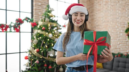 Téléchargez les photos : Jeune belle femme hispanique écoutant de la musique tenant cadeau célébrant Noël à la maison - en image libre de droit