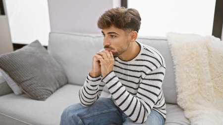 Photo for Worried young arab man with a beard, sitting on his living room sofa at home, seriously expressing doubt and sadness as he nervously ponders a problem indoors. - Royalty Free Image