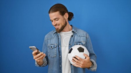 Photo for Young hispanic man smiling holding soccer ball and smartphone over isolated blue background - Royalty Free Image