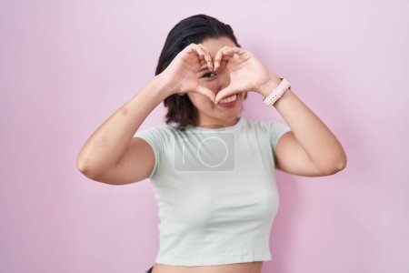 Photo for Hispanic young woman standing over pink background doing heart shape with hand and fingers smiling looking through sign - Royalty Free Image
