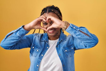 Téléchargez les photos : Femme afro-américaine avec des tresses debout sur fond jaune faisant forme de coeur avec la main et les doigts souriant en regardant à travers signe - en image libre de droit