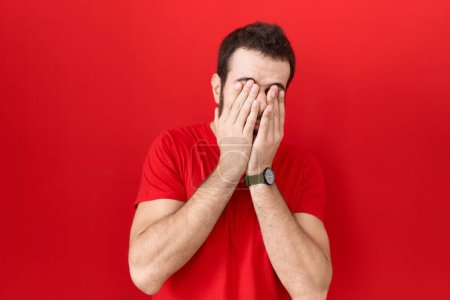 Foto de Hombre hispano joven con camiseta roja casual frotando los ojos para la fatiga y el dolor de cabeza, la expresión somnolienta y cansada. problema de visión - Imagen libre de derechos