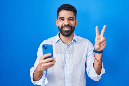 Photo for Hispanic man with beard using smartphone typing message smiling with happy face winking at the camera doing victory sign with fingers. number two. - Royalty Free Image