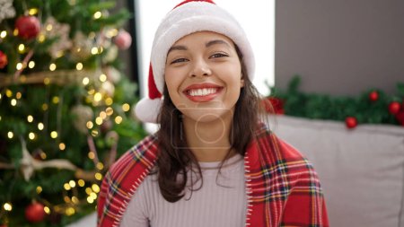 Photo for Young beautiful hispanic woman smiling sitting by christmas tree at home - Royalty Free Image