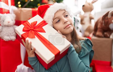 Téléchargez les photos : Adorable fille hispanique tenant cadeau assis sur le sol par l'arbre de Noël à la maison - en image libre de droit