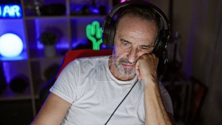 Foto de Hombre de mediana edad, pelo gris y cara aburrida, juegos de streaming en una habitación oscura, la vida de un jugador adulto - Imagen libre de derechos