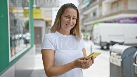 Téléchargez les photos : Belle femme blonde adulte utilisant joyeusement son smartphone en plein air, écran tactile, tapant un message texte joyeux dans la rue de la ville. - en image libre de droit