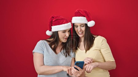 Téléchargez les photos : Deux femmes portant un chapeau de Noël en utilisant un smartphone sur fond rouge isolé - en image libre de droit