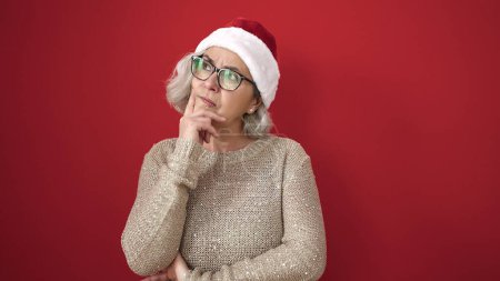 Téléchargez les photos : Femme d'âge moyen aux cheveux gris portant un chapeau de Noël avec une expression de doute sur fond rouge isolé - en image libre de droit