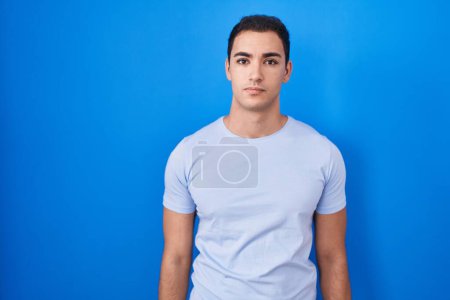 Photo for Young hispanic man standing over blue background relaxed with serious expression on face. simple and natural looking at the camera. - Royalty Free Image