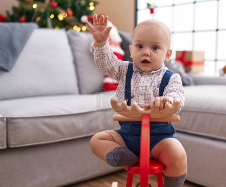 Photo for Adorable caucasian baby playing with reindeer rocking by christmas tree with hand raised up at home - Royalty Free Image