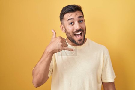 Photo for Handsome hispanic man standing over yellow background smiling doing phone gesture with hand and fingers like talking on the telephone. communicating concepts. - Royalty Free Image