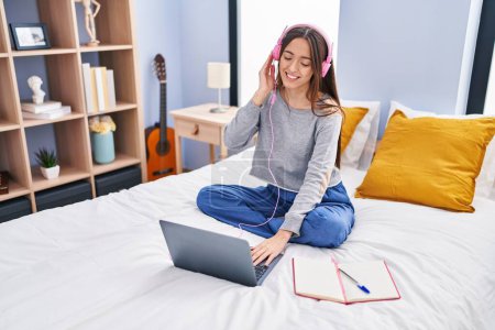Téléchargez les photos : Jeune belle étudiante hispanique écoutant de la musique vto étudier sur le lit dans la chambre - en image libre de droit
