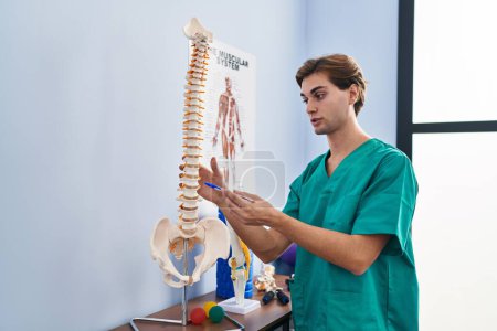 Téléchargez les photos : Jeune homme caucasien physiothérapeute touchant le modèle anatomique de la colonne vertébrale à la clinique de réadaptation - en image libre de droit