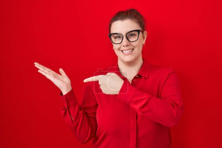 Foto de Mujer hispana joven con el pelo rojo de pie sobre el fondo rojo asombrado y sonriendo a la cámara mientras se presenta con la mano y señalando con el dedo. - Imagen libre de derechos