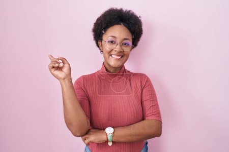 Téléchargez les photos : Belle femme africaine avec les cheveux bouclés debout sur fond rose avec un grand sourire sur le visage, pointant avec le doigt de la main vers le côté regardant la caméra. - en image libre de droit