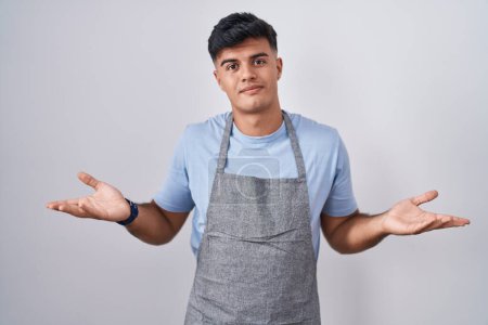 Photo for Hispanic young man wearing apron over white background clueless and confused with open arms, no idea concept. - Royalty Free Image