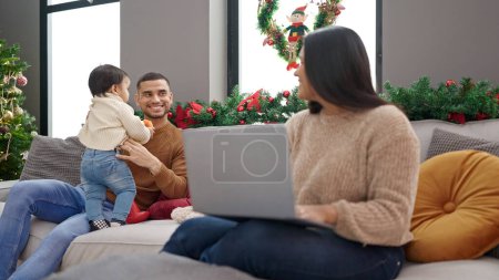 Téléchargez les photos : Couple et fils prenant soin de son fils assis sur le canapé par arbre de Noël travaillant à la maison - en image libre de droit