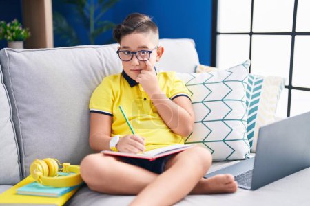 Photo for Young hispanic kid doing homework sitting on the sofa pointing to the eye watching you gesture, suspicious expression - Royalty Free Image