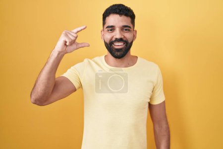 Téléchargez les photos : Homme hispanique avec barbe debout sur fond jaune geste souriant et confiant avec la main faisant signe de petite taille avec les doigts regardant et la caméra. concept de mesure. - en image libre de droit