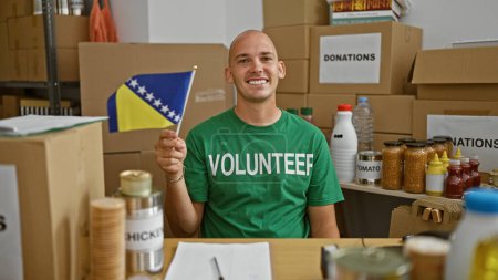 Foto de Joven hombre hispano voluntario sonriendo confiado sosteniendo bandera bosnia en el centro de caridad - Imagen libre de derechos